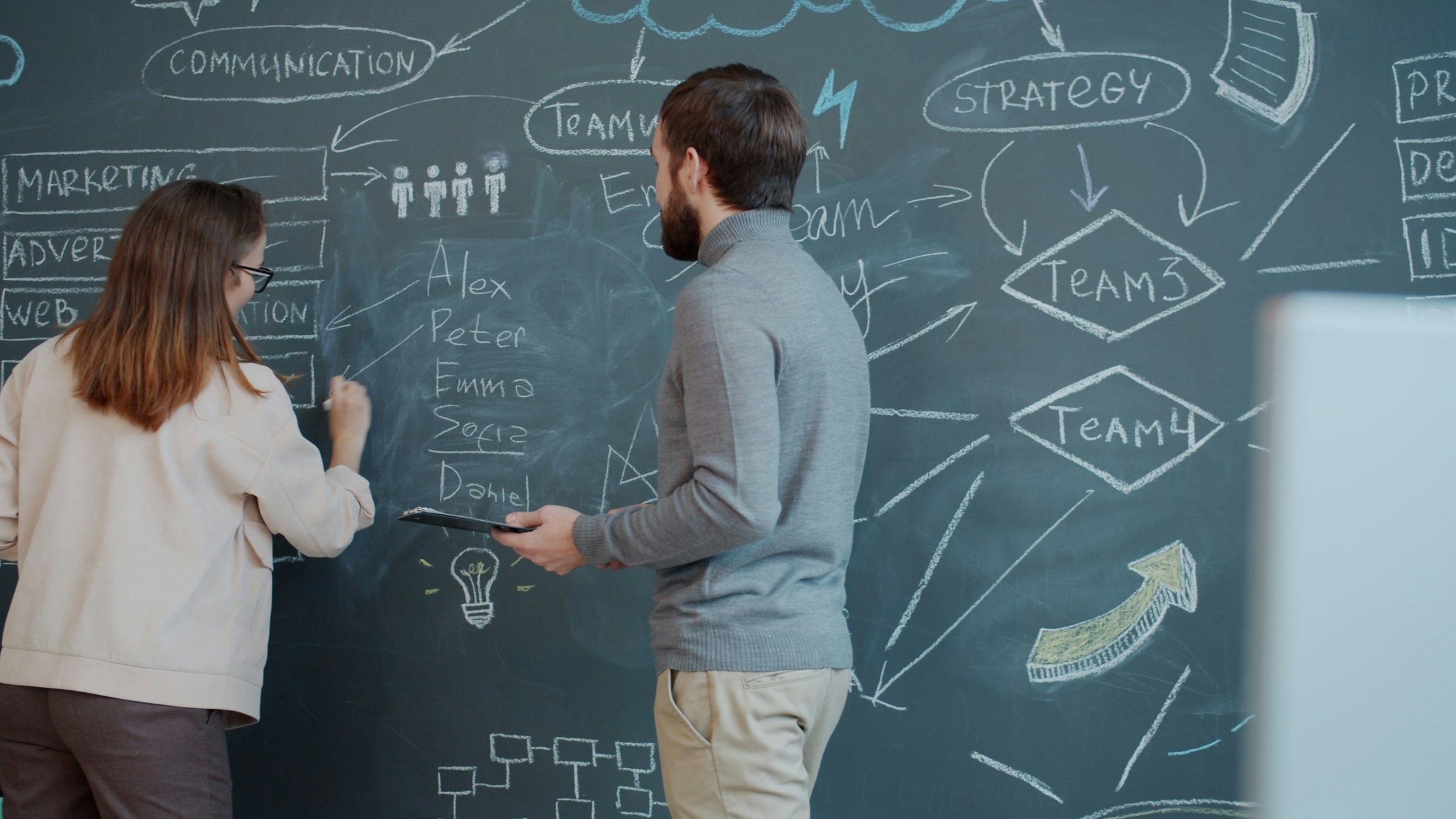 Two people mapping a strategy diagram in chalk on a wall — communication, marketing, team, and strategy nodes connected by arrows.