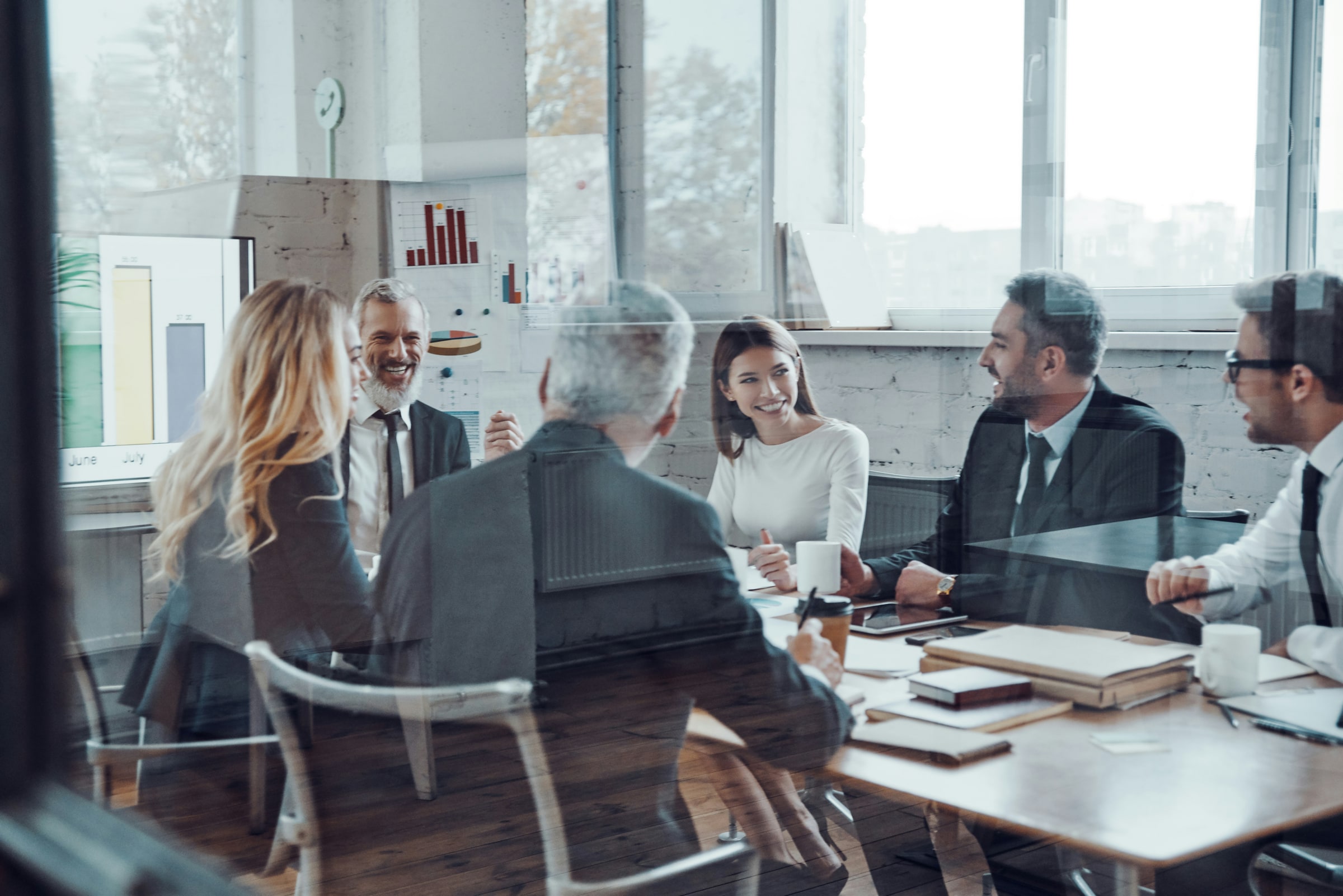 Business team in suits around a conference-room table in conversation, financial charts on the wall behind.